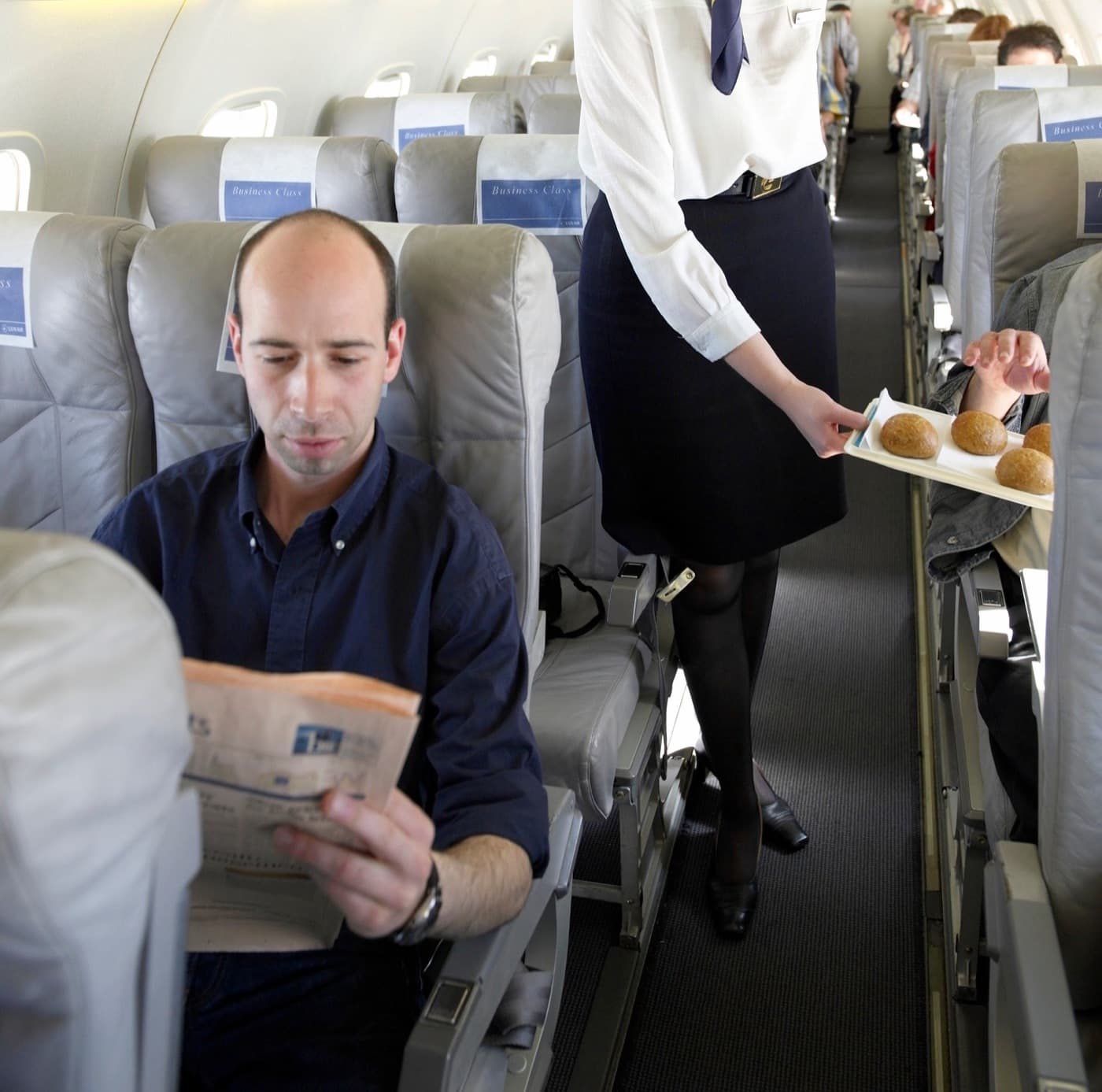 Letuška ponúka pečivo cestujúcim v business class, muž si číta noviny