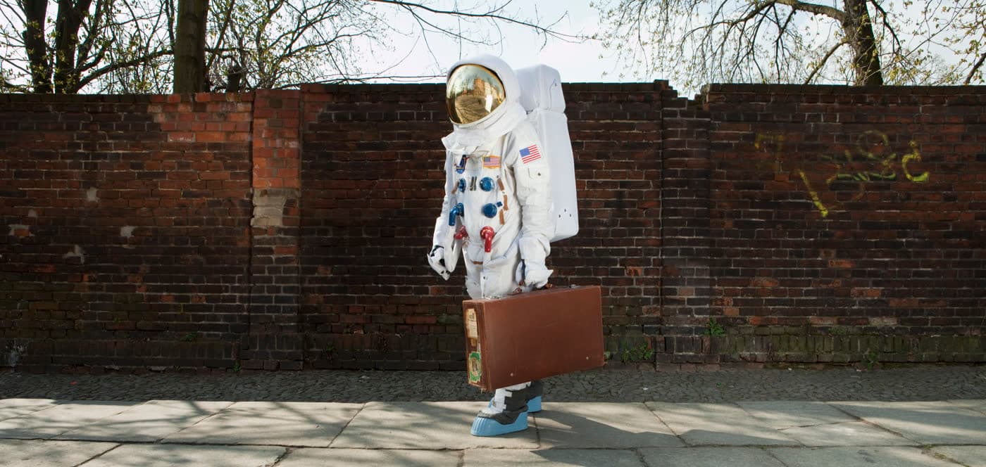 Astronaut s koženým kufrom v ruke čaká na chodníku pri tehlovej stene